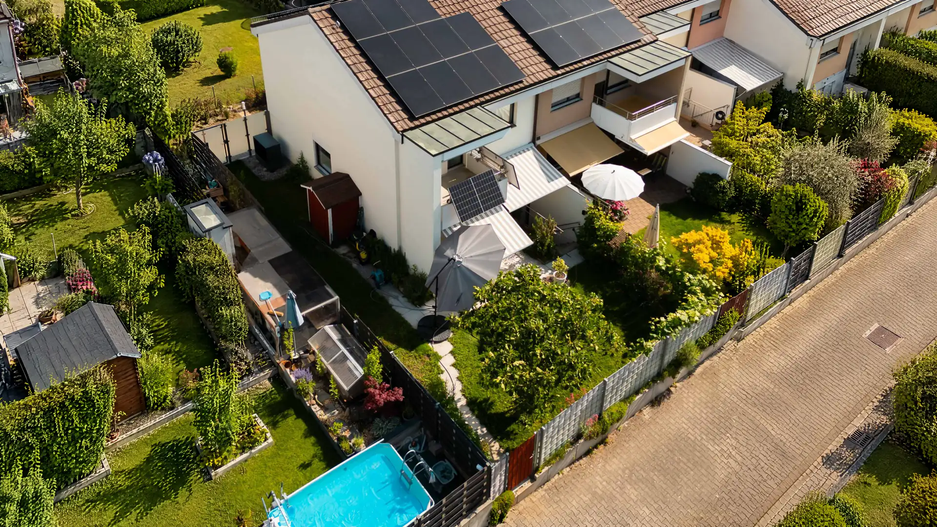 Reihenendhaus mit schwarzen Solarmodulen auf dem Ziegeldach und einem Solarmodul am Balkongeländer, Garten mit Sonnenschirmen, Drohnenblick