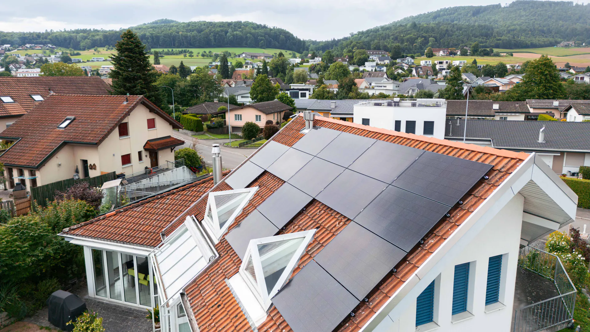 Rotes Ziegeldach mit schwarzen Solarmodulen neben zwei aufgestellten Dachfenstern, dahinter ein Dorf und bewaldete Hügel