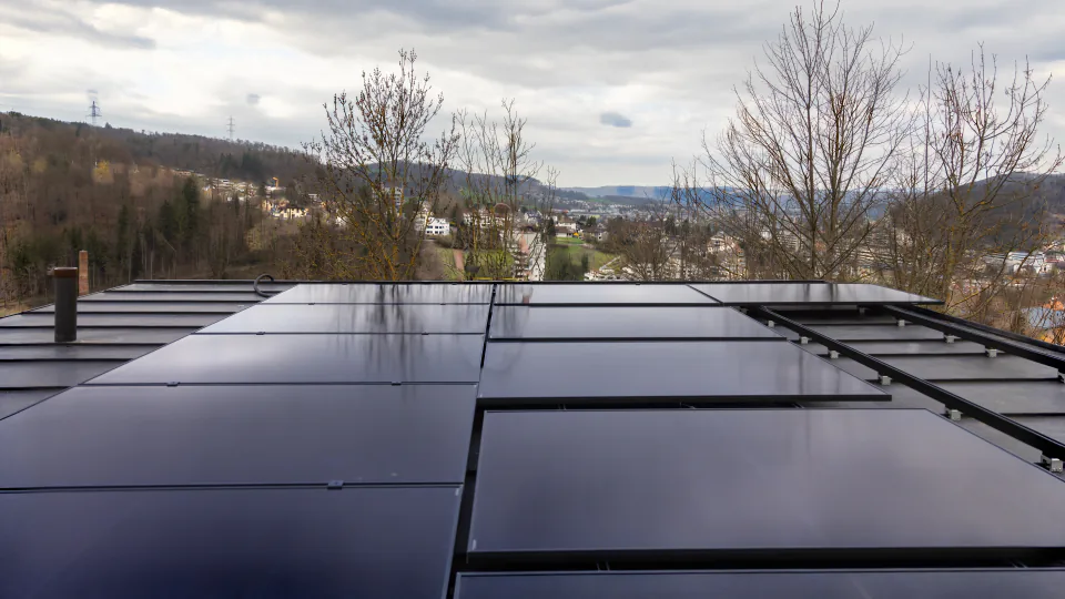 Flachdach mit Reihen schwarzer Solarmodule über einem Tal mit Wohnquartier, bewölkter Himmel
