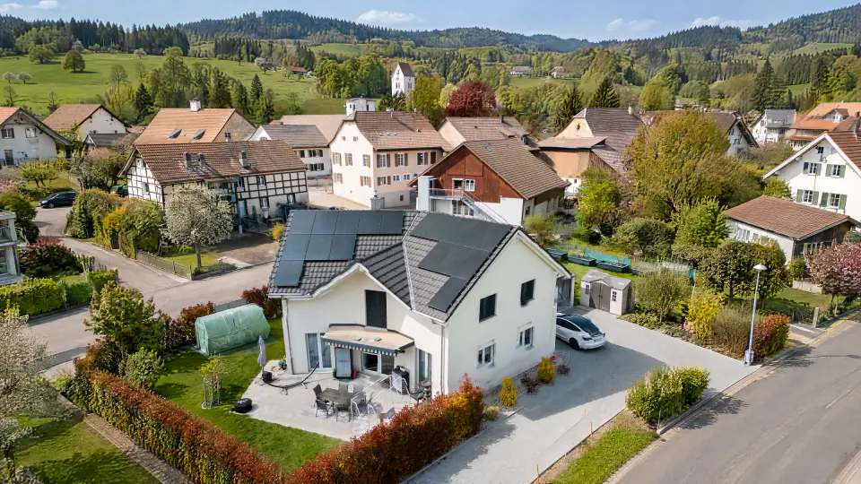 Weisses Einfamilienhaus mit schwarzen Solarmodulen auf beiden Dachseiten in einem Schweizer Dorf, Drohnenblick im Frühling