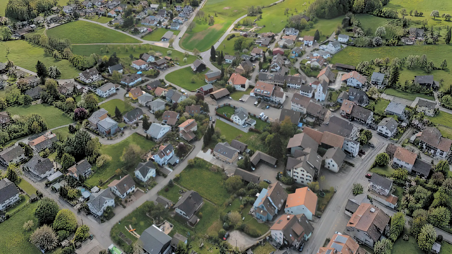 Dorf von oben mit Einfamilienhäusern, deren Satteldächer in alle Himmelsrichtungen zeigen, dazwischen Wiesen und Strassen